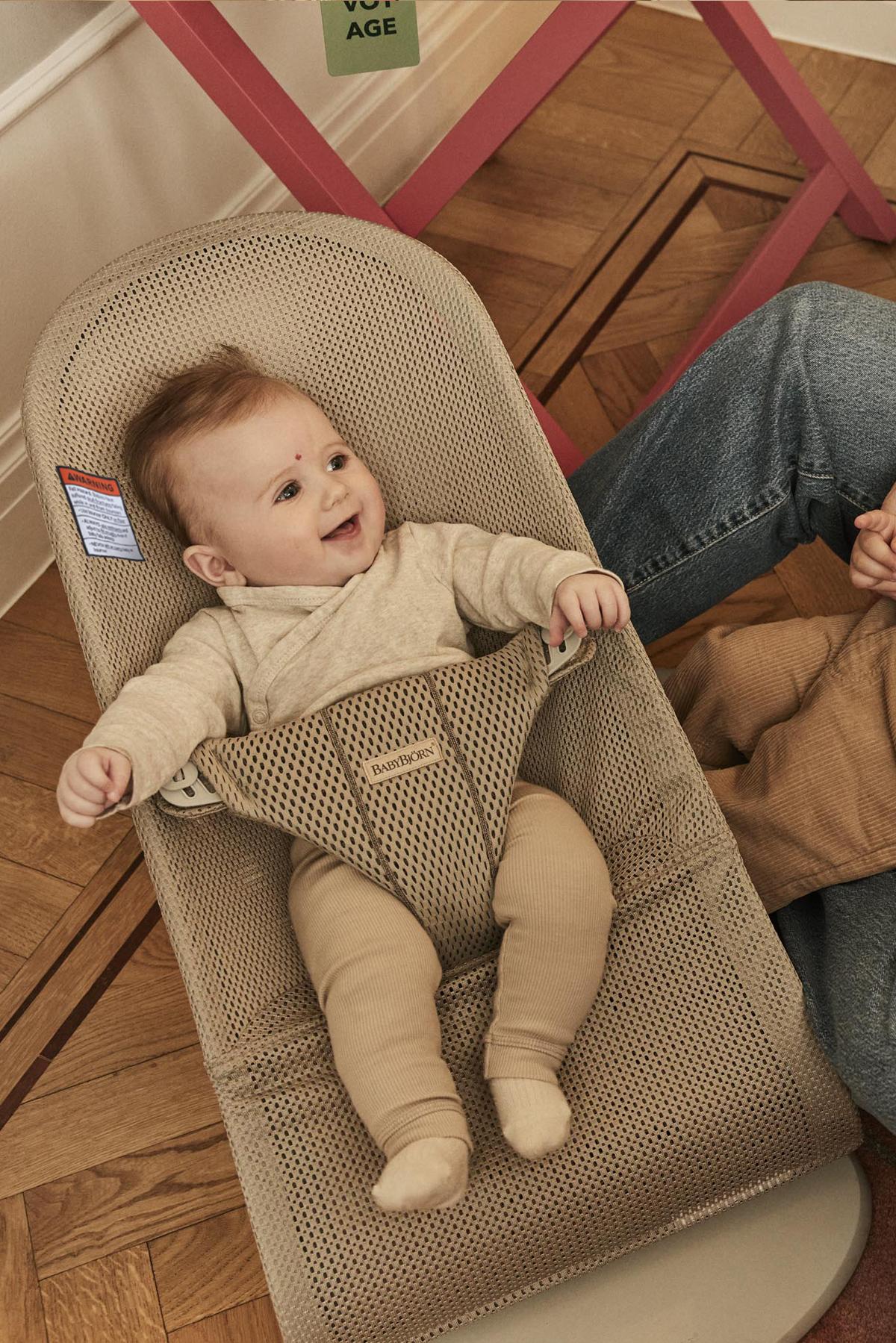 A smiling baby in a beige bouncer seat, wearing a light outfit, with an adult's arm visible nearby on a wooden floor.