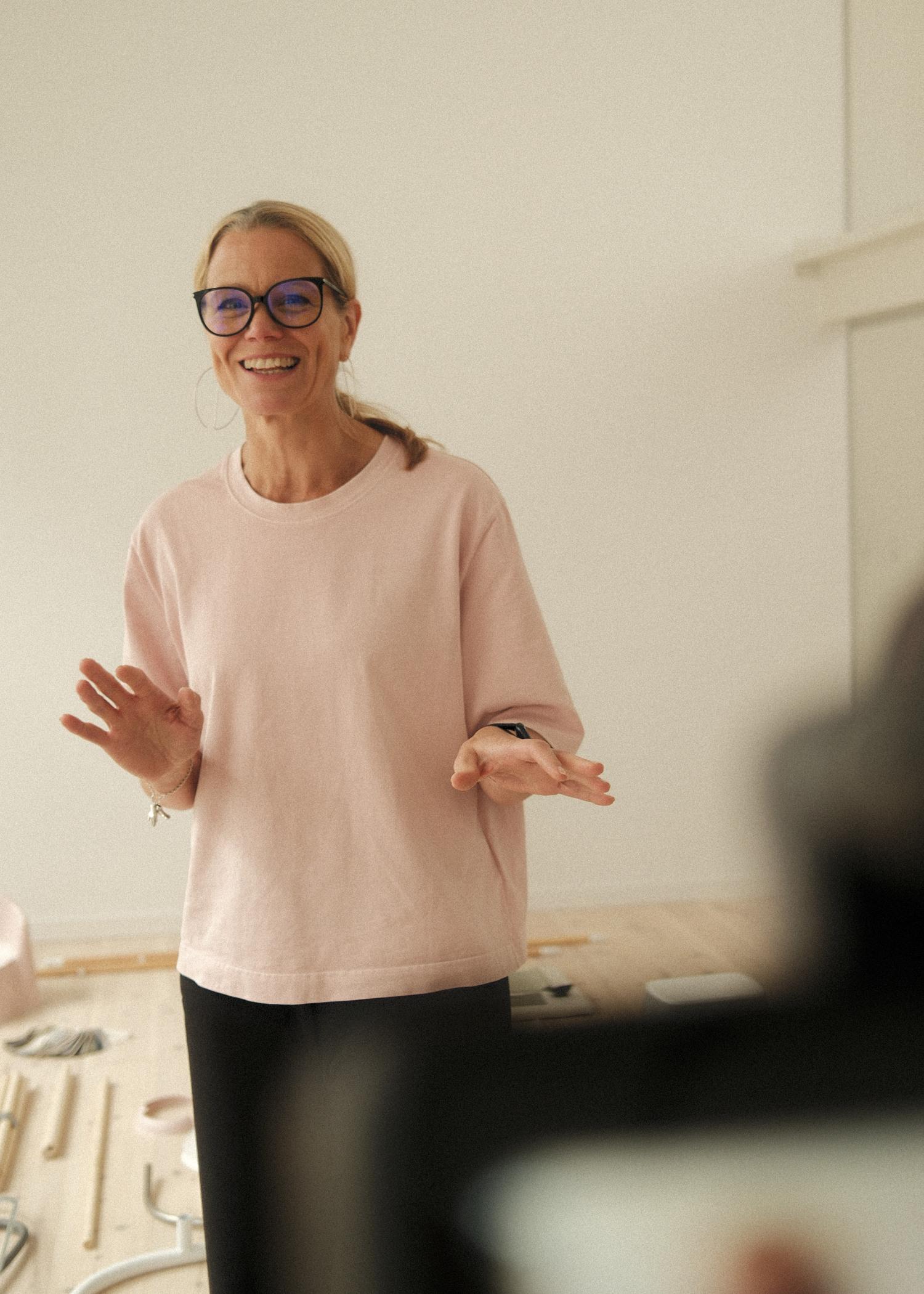 Smiling woman with glasses, wearing a pink shirt, gestures with hands in a bright room with scattered objects on the floor.