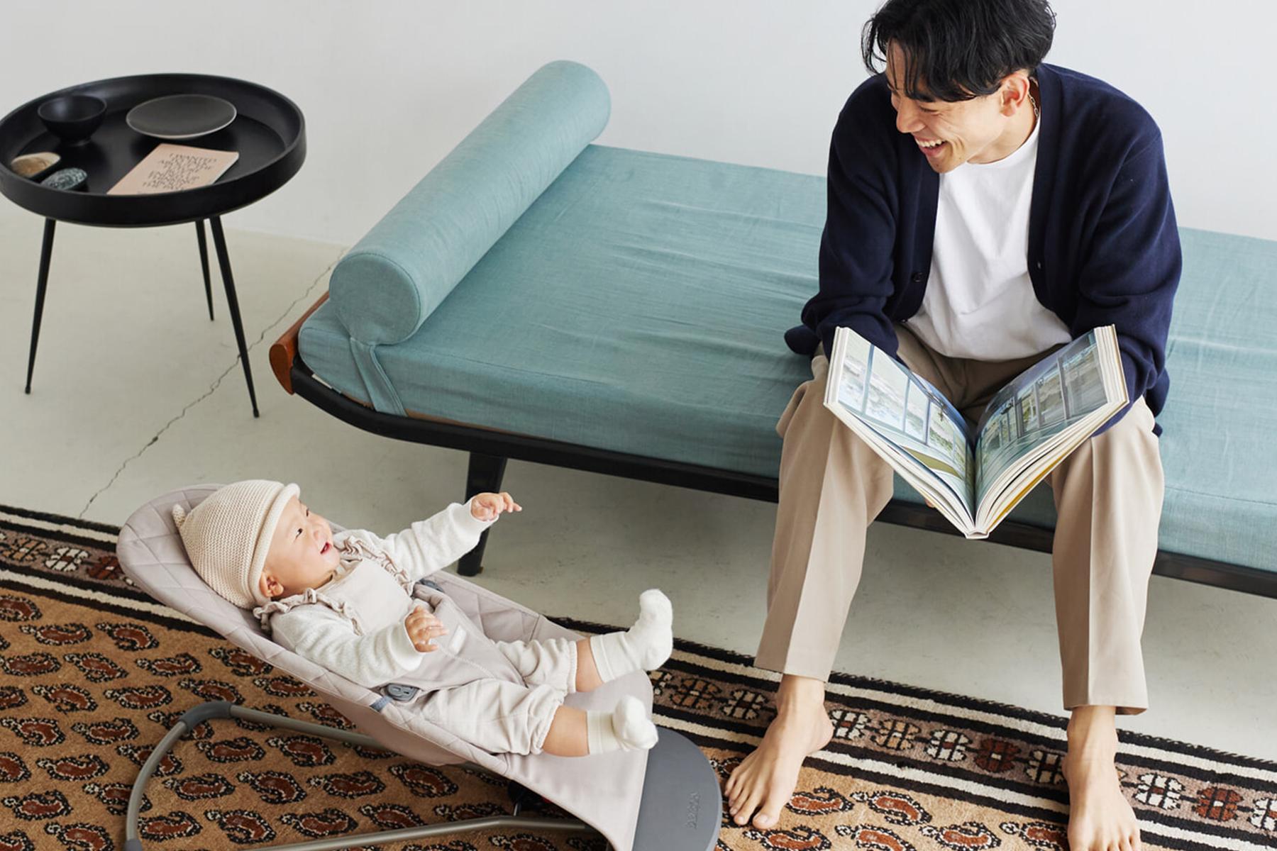 A man sits on a sofa, smiling and holding a book, while a baby in a bouncer looks up at him. A small round table is nearby.