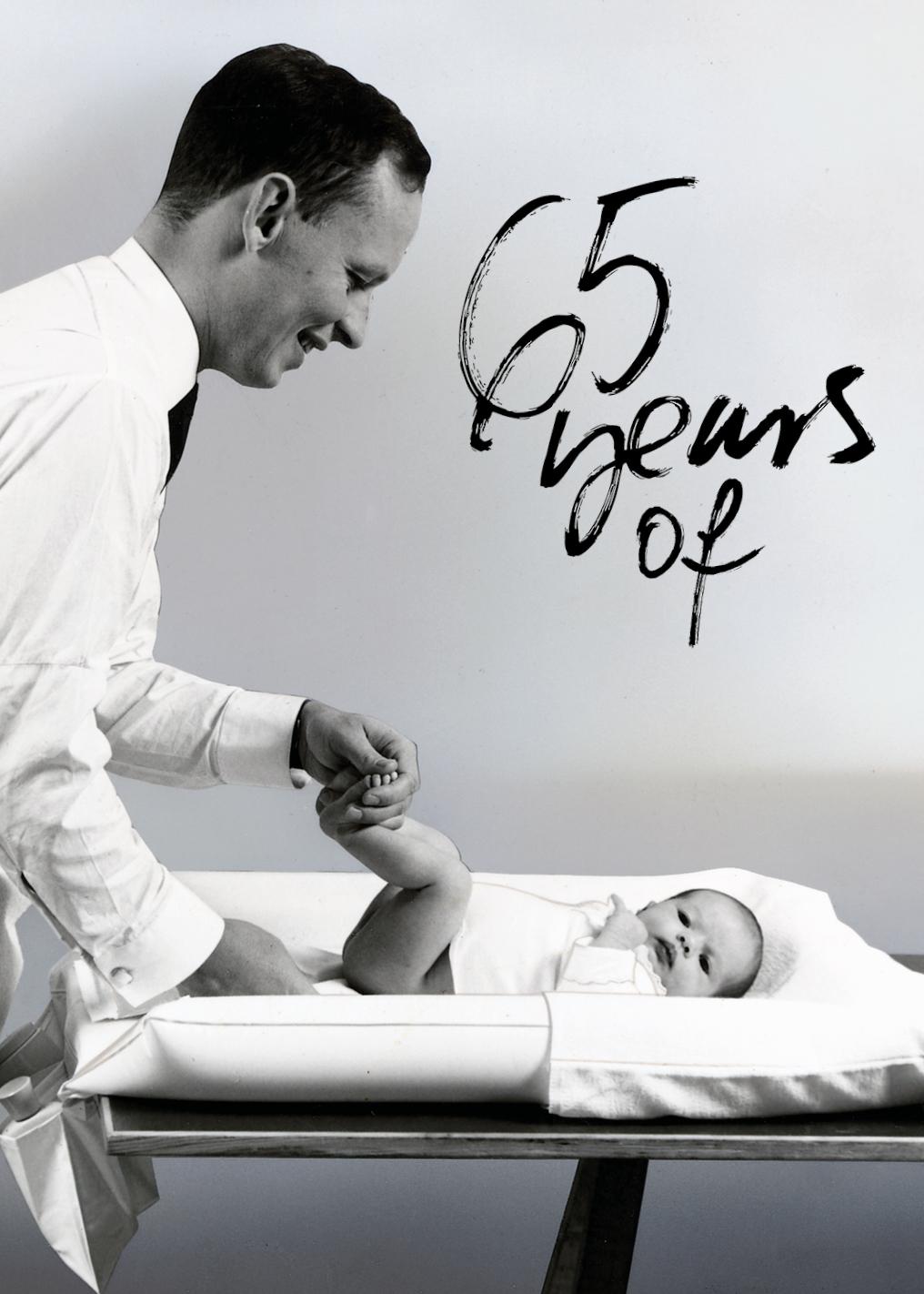 A man smiling at a baby on a changing table. Text reads "65 years of" in artistic script.