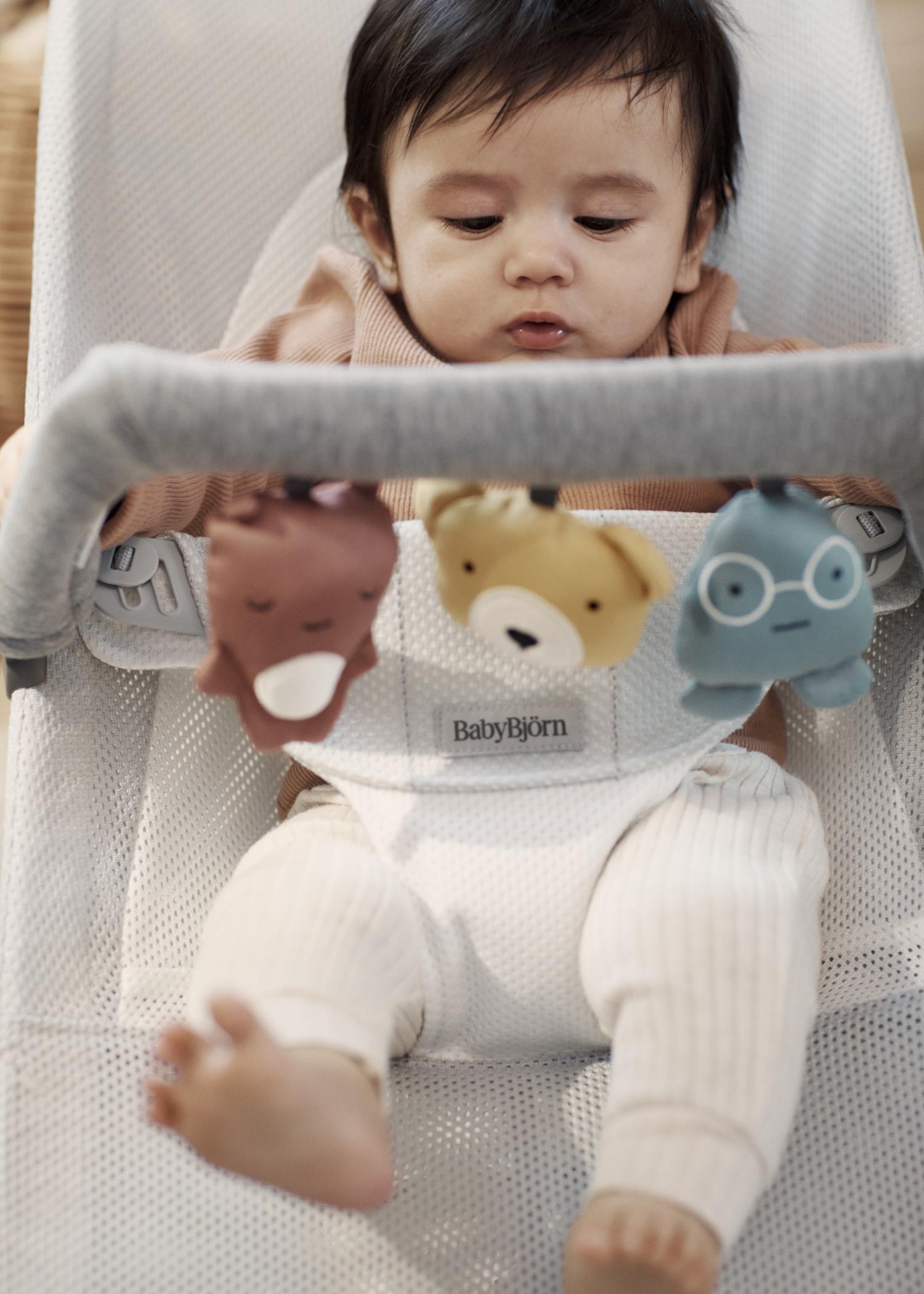 Baby seated in a bouncer, looking at hanging animal toys. The bouncer is white with a soft fabric, and the baby wears light-colored clothing.