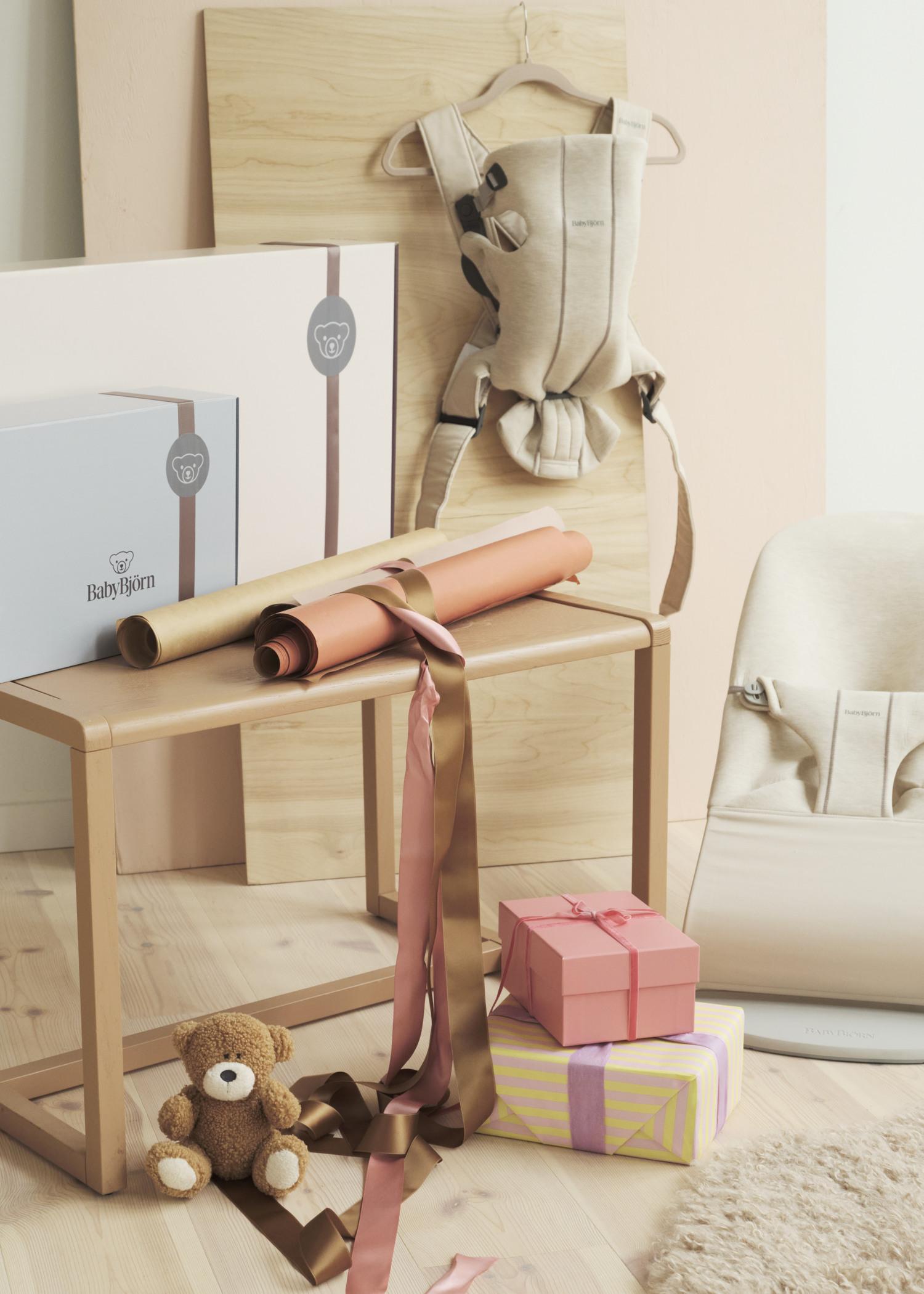 A baby carrier, gift boxes, wrapping paper, ribbons, a teddy bear, and a baby seat are arranged on and around a wooden table.
