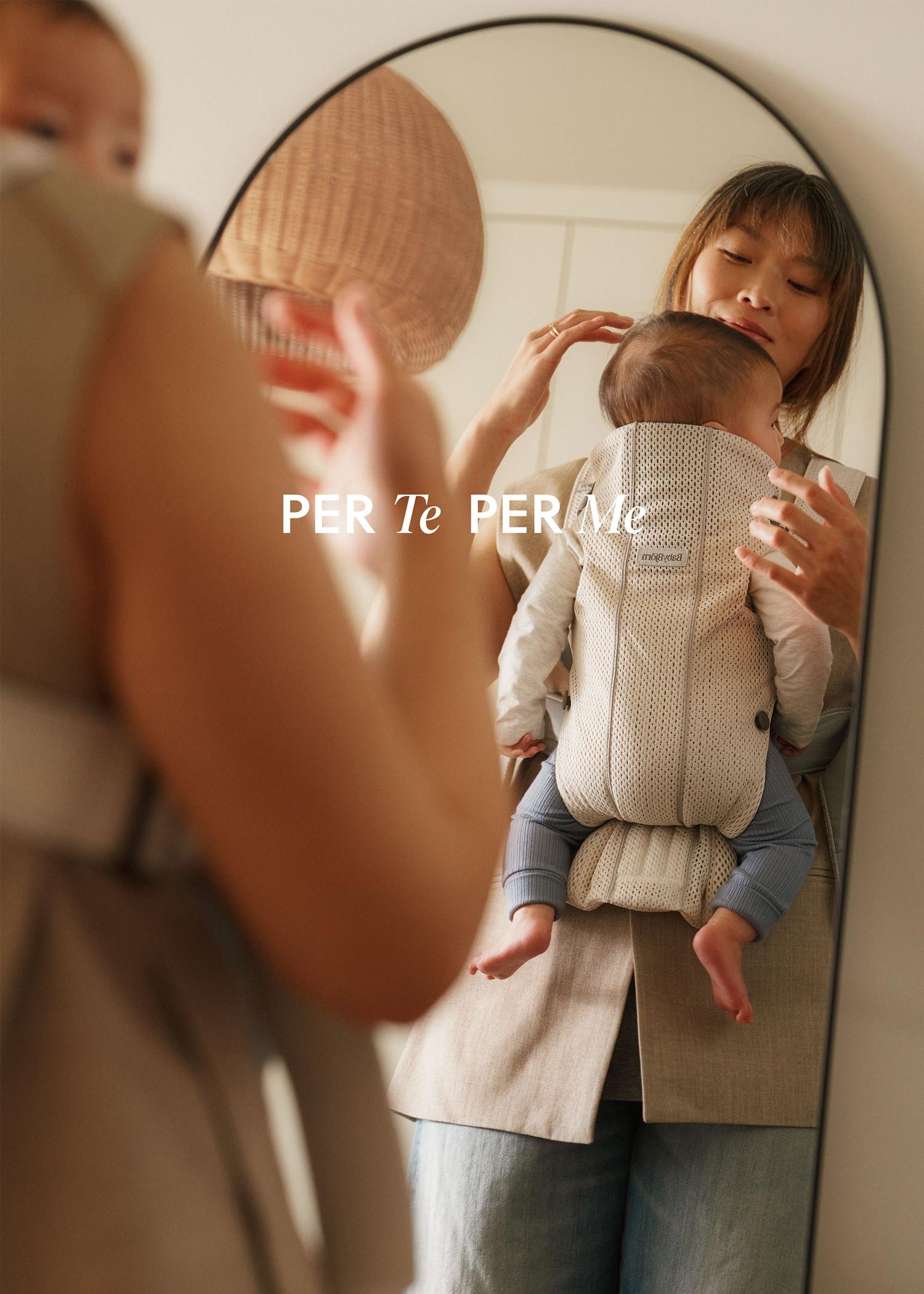 Woman carrying a baby in a light-colored carrier, looking in a mirror. Another person in a hat is visible in the foreground.