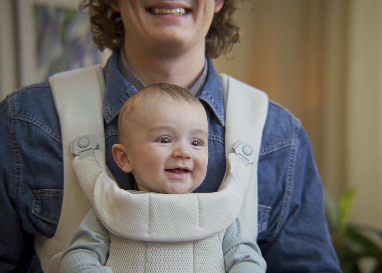 A smiling baby in a white carrier worn by a person in a denim shirt.