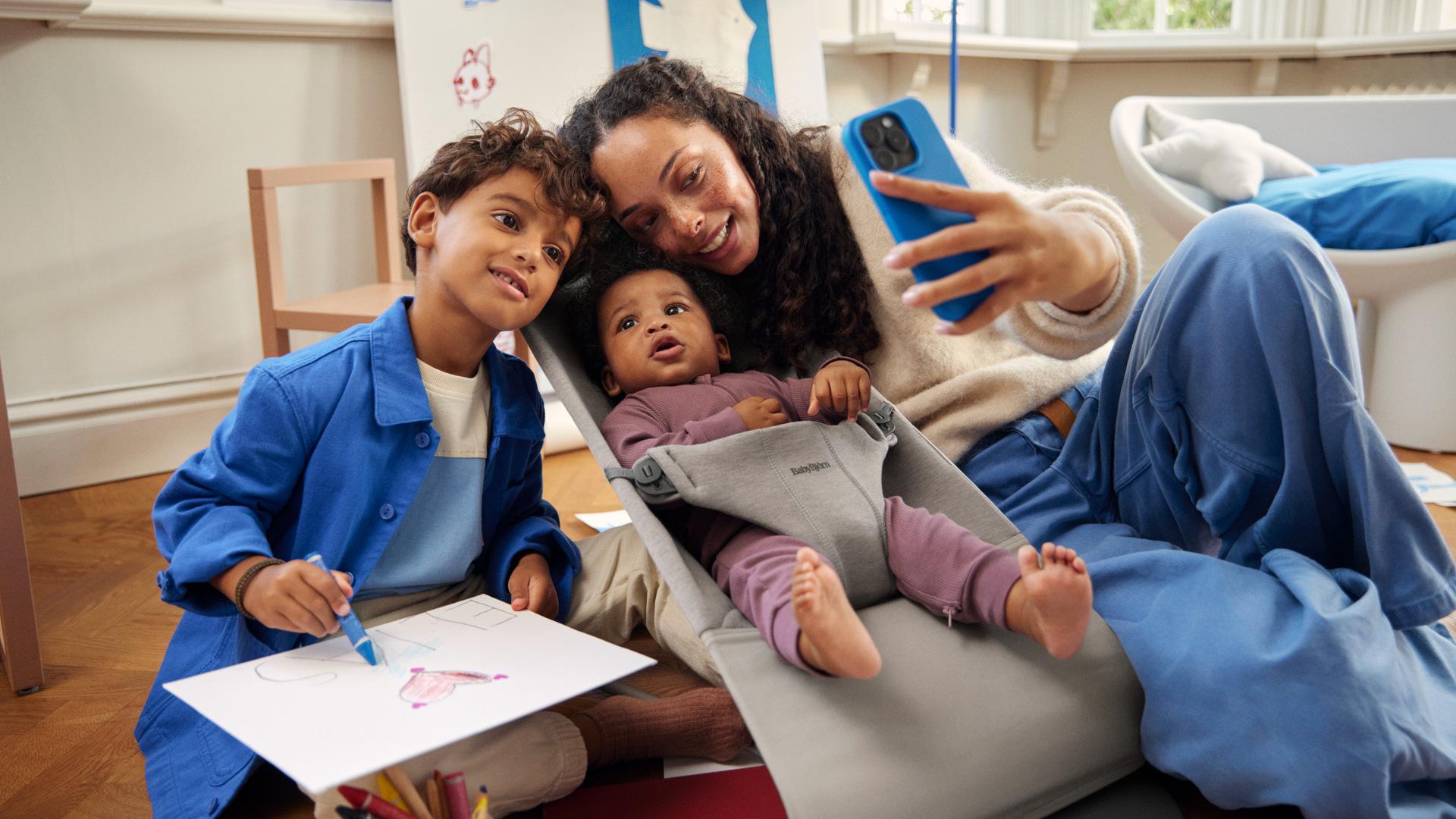 A woman takes a selfie with two children; one is drawing, and the other is in a baby bouncer. They are in a cozy room.