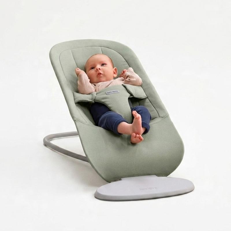 A baby sits comfortably in a light green baby bouncer against a plain background, wearing a light sweater and dark pants.